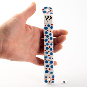 May include: White ceramic mezuzah with a blue and red polka dot pattern. The mezuzah has a Hebrew letter Shin on the front.