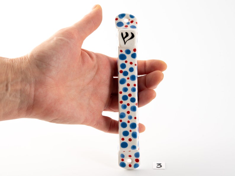 May include: White ceramic mezuzah with a blue and red polka dot pattern. The mezuzah has a Hebrew letter Shin on the front.