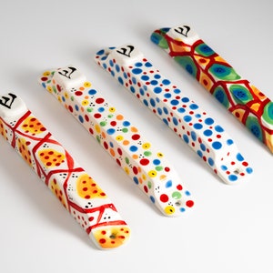 May include: Four colorful ceramic mezuzahs with different patterns. Each mezuzah has a white background with red, yellow, blue, and green accents. The mezuzahs are decorated with dots, geometric shapes, and abstract designs.