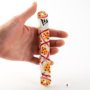 May include: A white mezuzah with a red and yellow geometric pattern. The mezuzah has the Hebrew letter "Shin" on the top.