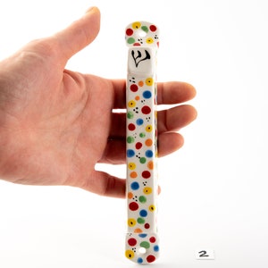 May include: A white ceramic mezuzah with a colorful polka dot pattern in red, yellow, blue, and green. The mezuzah has a Hebrew letter Shin on the front.