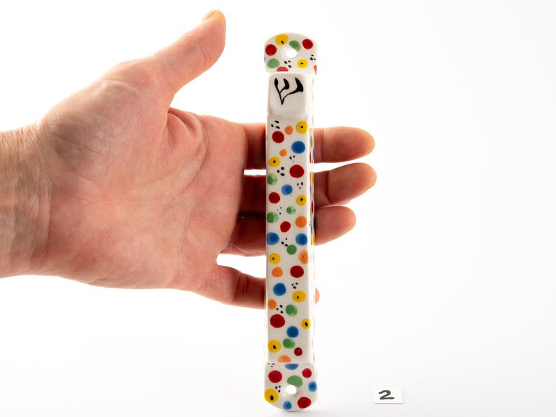 May include: A white ceramic mezuzah with a colorful polka dot pattern in red, yellow, blue, and green. The mezuzah has a Hebrew letter Shin on the front.