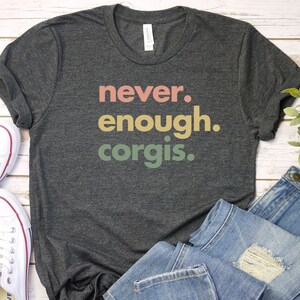 May include: A dark gray t-shirt with the text "never. enough. corgis." printed in colorful letters. The shirt is laid out on a white wooden surface with a pair of white sneakers and blue jeans.