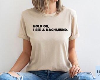 May include: A beige t-shirt with the text "Hold on, I see a dachshund." printed in black.