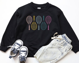 May include: Black sweatshirt with a graphic of five colorful tennis rackets. The sweatshirt is paired with light blue denim shorts and blue sneakers.