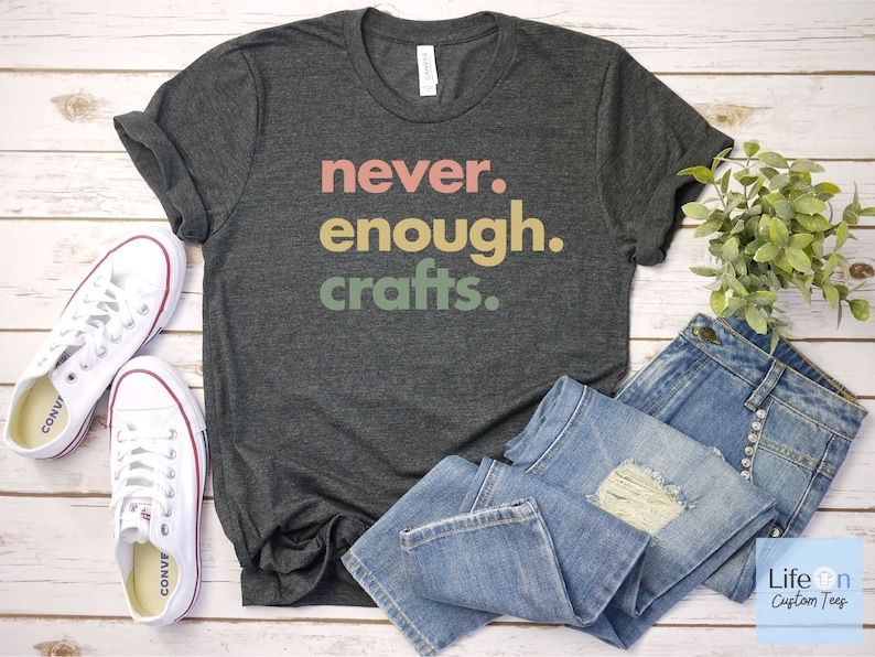 May include: A dark grey t-shirt with the text "never. enough. crafts." in colourful letters. The shirt is laid out on a white wooden surface with a pair of white trainers and blue jeans.