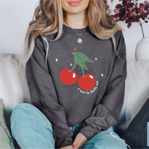 May include: A dark gray sweatshirt with a graphic of two red cherries with green stems and leaves. The text "c'est la belle vie" is printed below the cherries.