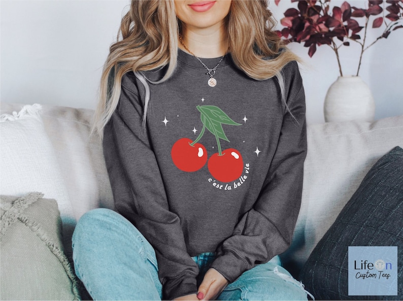 May include: A dark gray sweatshirt with a graphic of two red cherries with green stems and leaves. The text "c'est la belle vie" is printed below the cherries.