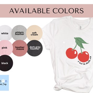 May include: A white t-shirt with a graphic of two red cherries and the text "c'est la belle vie". The shirt is available in ten colors: white, athletic heather, soft cream, pink, heather mauve, dark grey heather, black, navy, light pink, and light blue.