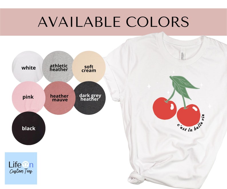 May include: A white t-shirt with a graphic of two red cherries and the text "c'est la belle vie". The shirt is available in ten colors: white, athletic heather, soft cream, pink, heather mauve, dark grey heather, black, navy, light pink, and light blue.