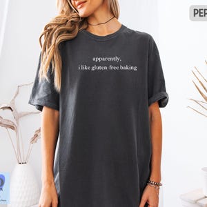 May include: Dark gray t-shirt with the text "apparently, i like gluten-free baking" in white. The shirt is a relaxed fit and has rolled-up sleeves. The word "PEPPER" is in a gray rectangle in the upper right corner.