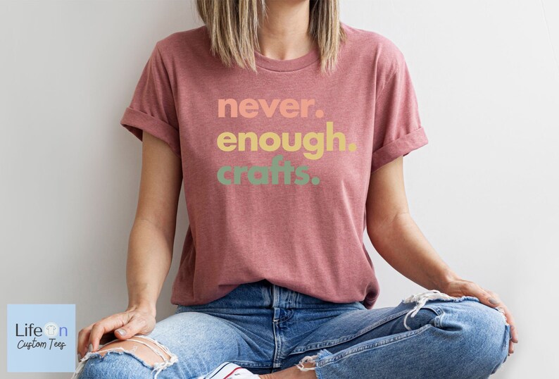 May include: A woman wearing a maroon t-shirt with the text "never. enough. crafts." in a gradient of peach, yellow, and green. She is sitting with her legs crossed and wearing blue jeans.