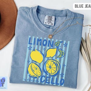 May include: A blue jean colored t-shirt with a lemon graphic. The graphic includes the words "LIMON CELLO" and "la dolce vita" with illustrations of lemons. A gold necklace is draped across the shirt.