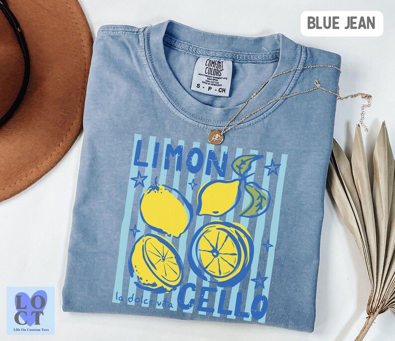 May include: A blue jean colored t-shirt with a lemon graphic. The graphic includes the words "LIMON CELLO" and "la dolce vita" with illustrations of lemons. A gold necklace is draped across the shirt.