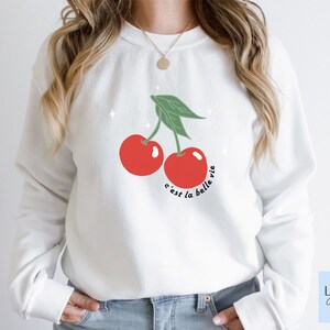 May include: White sweatshirt with a red cherry graphic and the text "c'est la belle vie" in black.
