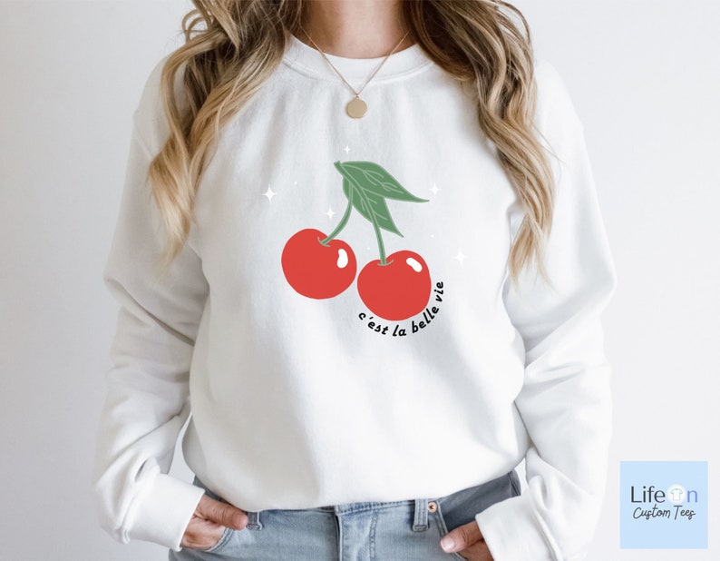 May include: White sweatshirt with a red cherry graphic and the text "c'est la belle vie" in black.