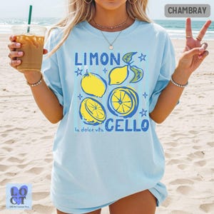 May include: A light blue t-shirt featuring a graphic print of yellow lemons and blue leaves with the text LIMON CELLO and la dolce vita. The design includes star accents and a circular lemon slice illustration on the front of the garment.