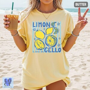 May include: A light yellow t-shirt with a graphic design featuring lemons, the words "LIMONCELLO" and "la dolce vita" in blue. The shirt is a relaxed fit, perfect for casual wear. The word "BUTTER" is printed in the top right corner.