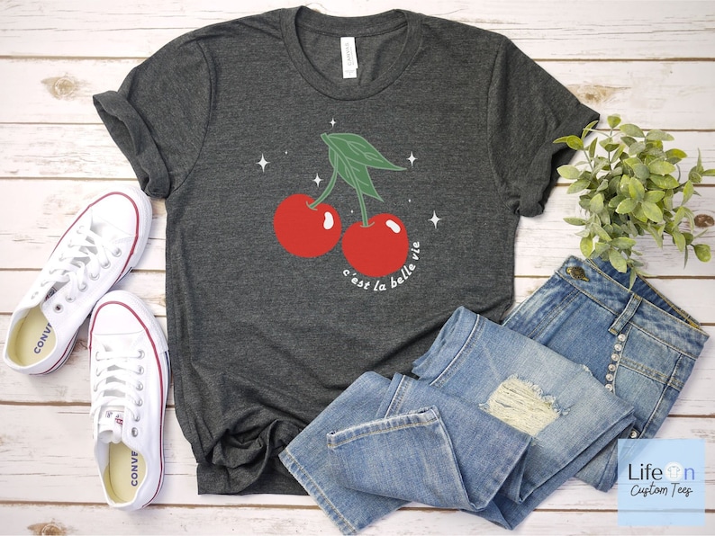 May include: A dark gray t-shirt with a graphic of two red cherries with green stems and leaves. The text "c'est la belle vie" is printed below the cherries.
