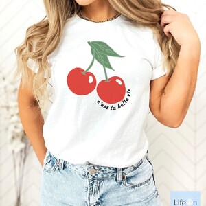 May include: White t-shirt with a graphic of two red cherries and the text "c'est la belle vie" in black.