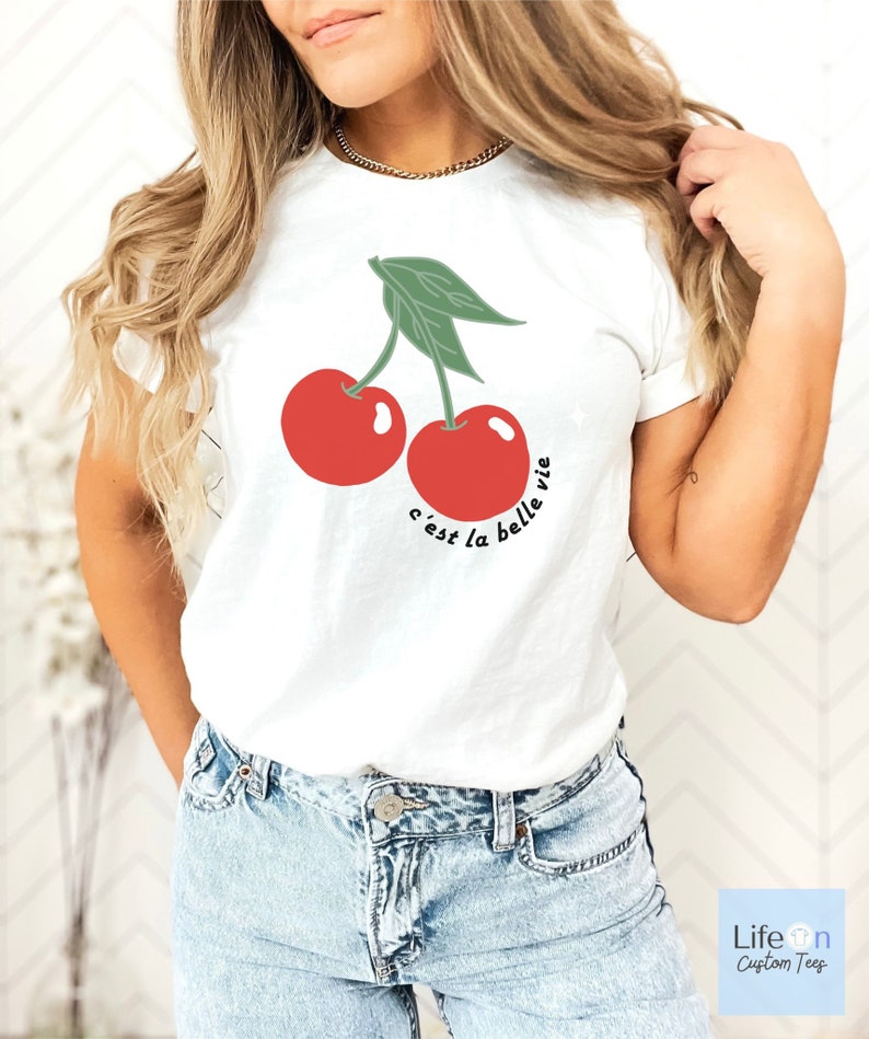 May include: White t-shirt with a graphic of two red cherries and the text "c'est la belle vie" in black.