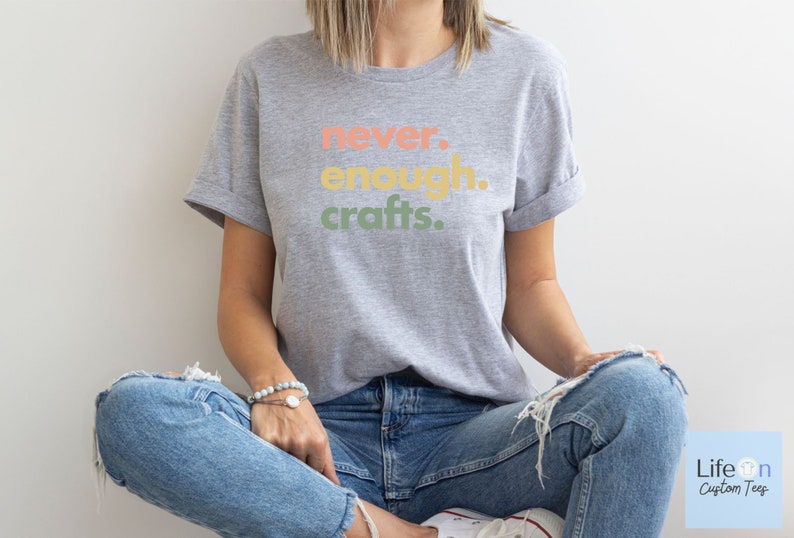 May include: A grey t-shirt with the text "never. enough. crafts." in colourful letters. The shirt is being worn by a person sitting with their legs crossed.