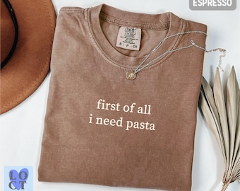 May include: Espresso-colored t-shirt with the text "first of all i need pasta" in white. The shirt has a crew neck and a Comfort Colors tag. A silver necklace with a pendant is draped across the shirt.