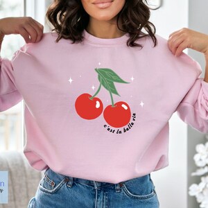 May include: Pink sweatshirt with a graphic of two red cherries and green leaves. The text "c'est la belle vie" is written below the cherries.