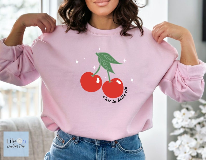 May include: Pink sweatshirt with a graphic of two red cherries and green leaves. The text "c'est la belle vie" is written below the cherries.
