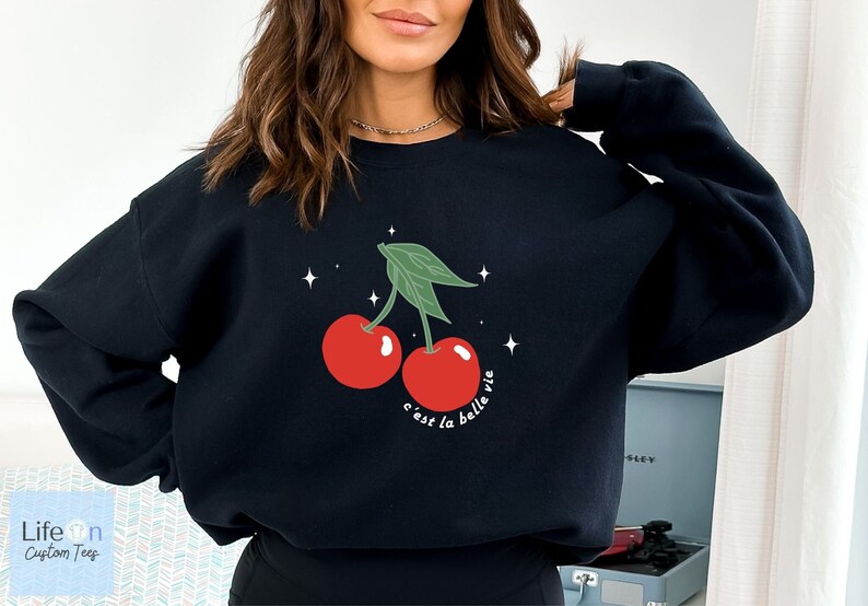 May include: Black sweatshirt with a graphic of two red cherries with green stems and leaves. The text "c'est la belle vie" is printed below the cherries.