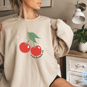 May include: A beige sweatshirt with a graphic of two red cherries and green leaves. The text "c'est la belle vie" is printed below the cherries.