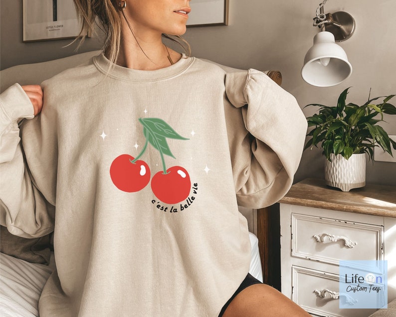 May include: A beige sweatshirt with a graphic of two red cherries and green leaves. The text "c'est la belle vie" is printed below the cherries.