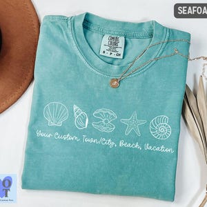 May include: Seafoam green t-shirt with white seashell graphics and the text "Your Custom Town/City, Beach, Vacation." A gold necklace with a pendant rests on the shirt. The shirt is folded next to a brown hat and dried leaves.