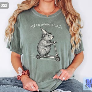 Puede incluir: Camiseta verde musgo con la frase "Off to avoid emails". Un dise&ntilde;o detallado de un roedor con sombrero de mago montado en un patinete est&aacute; impreso en la parte delantera. La camiseta es de estilo casual y c&oacute;modo.