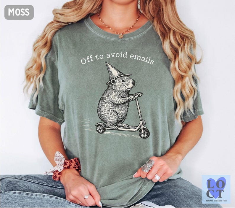 Puede incluir: Camiseta verde musgo con la frase "Off to avoid emails". Un dise&ntilde;o detallado de un roedor con sombrero de mago montado en un patinete est&aacute; impreso en la parte delantera. La camiseta es de estilo casual y c&oacute;modo.