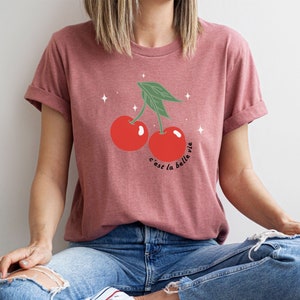 May include: A maroon t-shirt with a graphic of two red cherries with green stems and leaves. The text "c'est la belle vie" is printed below the cherries.