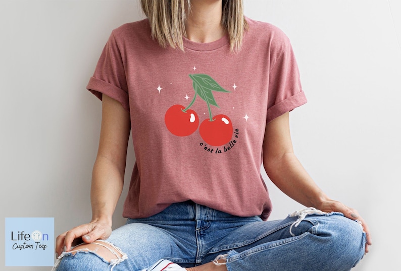 May include: A maroon t-shirt with a graphic of two red cherries with green stems and leaves. The text "c'est la belle vie" is printed below the cherries.