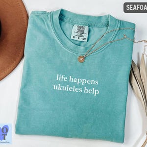 May include: Seafoam green t-shirt with the text "life happens ukuleles help" in white. A gold necklace with a pendant rests on the shirt. The shirt is a Comfort Colors brand.