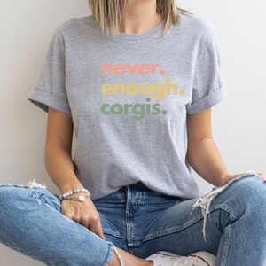 May include: A gray t-shirt with the text "never. enough. corgis." printed in colorful letters. The shirt is being worn by a person sitting with their legs crossed.