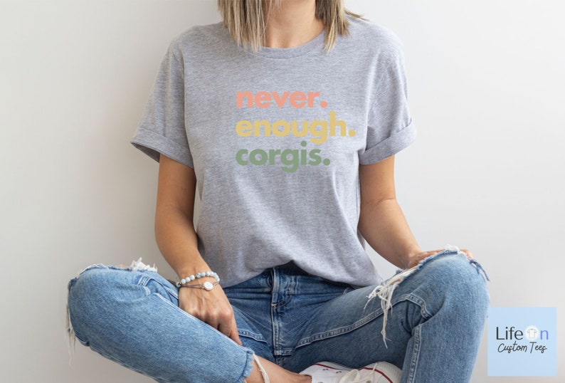 May include: A gray t-shirt with the text "never. enough. corgis." printed in colorful letters. The shirt is being worn by a person sitting with their legs crossed.