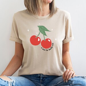 May include: A beige t-shirt with a graphic of two red cherries with green stems and leaves. The text "c'est la belle vie" is printed below the cherries.