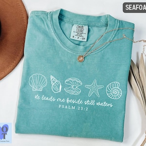 May include: Seafoam green t-shirt with white seashell graphics and the text "He leads me beside still waters, Psalm 23:2." The shirt has a crew neck and short sleeves. A gold necklace with a pendant is on the shirt.