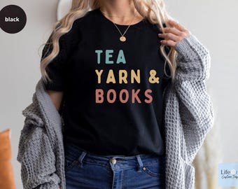 May include: A black t-shirt with the text "TEA YARN & BOOKS" in a retro color palette. The text is stacked vertically with the word "TEA" at the top and "BOOKS" at the bottom.
