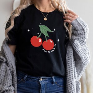 May include: Black t-shirt with a graphic of two red cherries with green stems and leaves. The text "c'est la belle vie" is printed below the cherries.