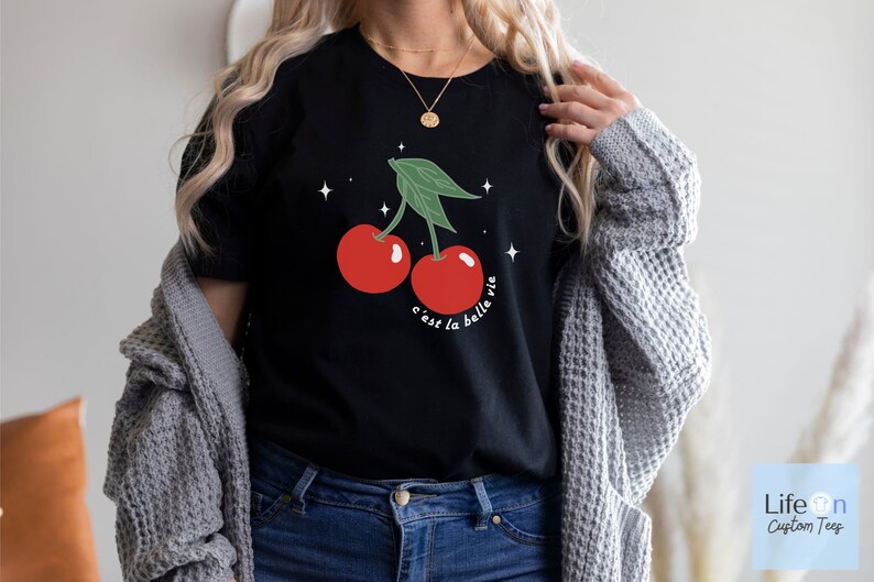 May include: Black t-shirt with a graphic of two red cherries with green stems and leaves. The text "c'est la belle vie" is printed below the cherries.