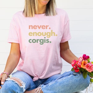 May include: A pink t-shirt with the text "never. enough. corgis." printed in a gradient of peach, yellow, and green.