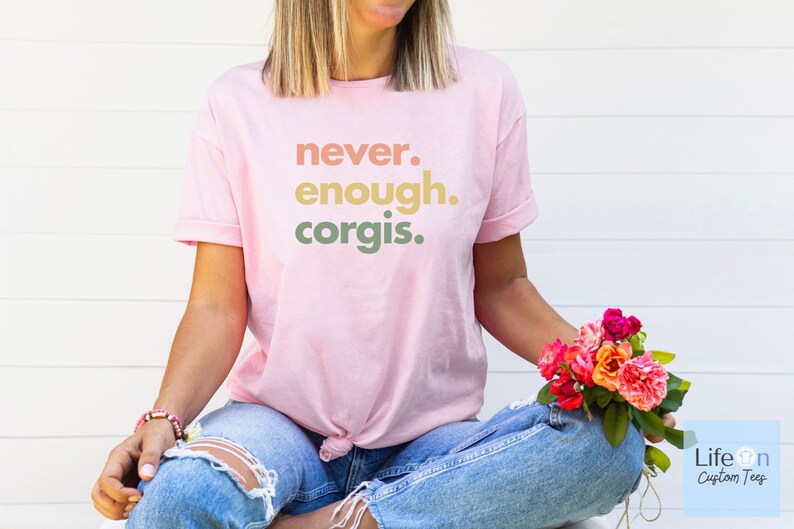 May include: A pink t-shirt with the text "never. enough. corgis." printed in a gradient of peach, yellow, and green.