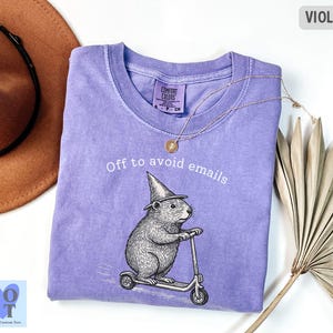 Puede incluir: Camiseta violeta con la frase "Off to avoid emails" y un gr&aacute;fico de un roedor en un scooter con un sombrero de mago. La camiseta tiene cuello redondo y mangas cortas. Un collar dorado est&aacute; drapeado sobre la camiseta.