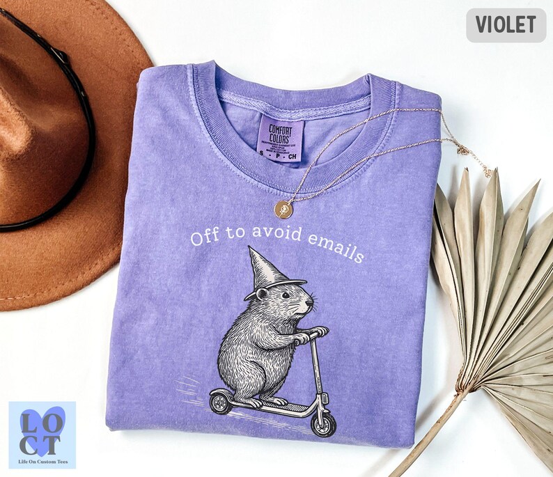 Puede incluir: Camiseta violeta con la frase "Off to avoid emails" y un gr&aacute;fico de un roedor en un scooter con un sombrero de mago. La camiseta tiene cuello redondo y mangas cortas. Un collar dorado est&aacute; drapeado sobre la camiseta.