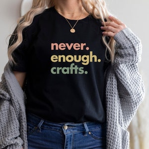 May include: A black t-shirt with the text "never. enough. crafts." in colourful lettering. The shirt is being worn by a person with long blonde hair and a grey cardigan.